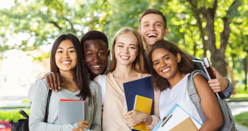 Group of international happy students with books and notebooks having fun in park after studying, smiling at camera